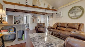 Una sala de estar acogedora con un sofá de cuero marrón, una mesita de centro y una alfombra. Se observa una zona de cocina con un refrigerador y un lavamanos.