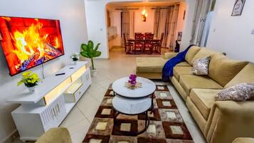 Sala de estar com televisão de tela plana, sofá, mesa de centro e área de jantar ao fundo.