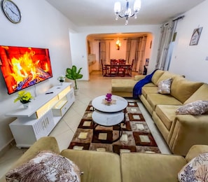 Sala de estar com televisão de tela plana, sofá, mesa de centro e área de jantar ao fundo.