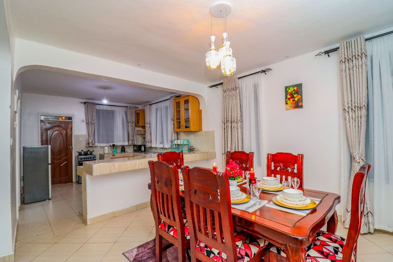 Uma área de refeições com mesa de madeira posta para quatro pessoas, uma cozinha ao fundo e um candelabro acima.