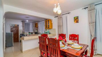 Uma área de refeições com mesa de madeira posta para quatro pessoas, uma cozinha ao fundo e um candelabro acima.
