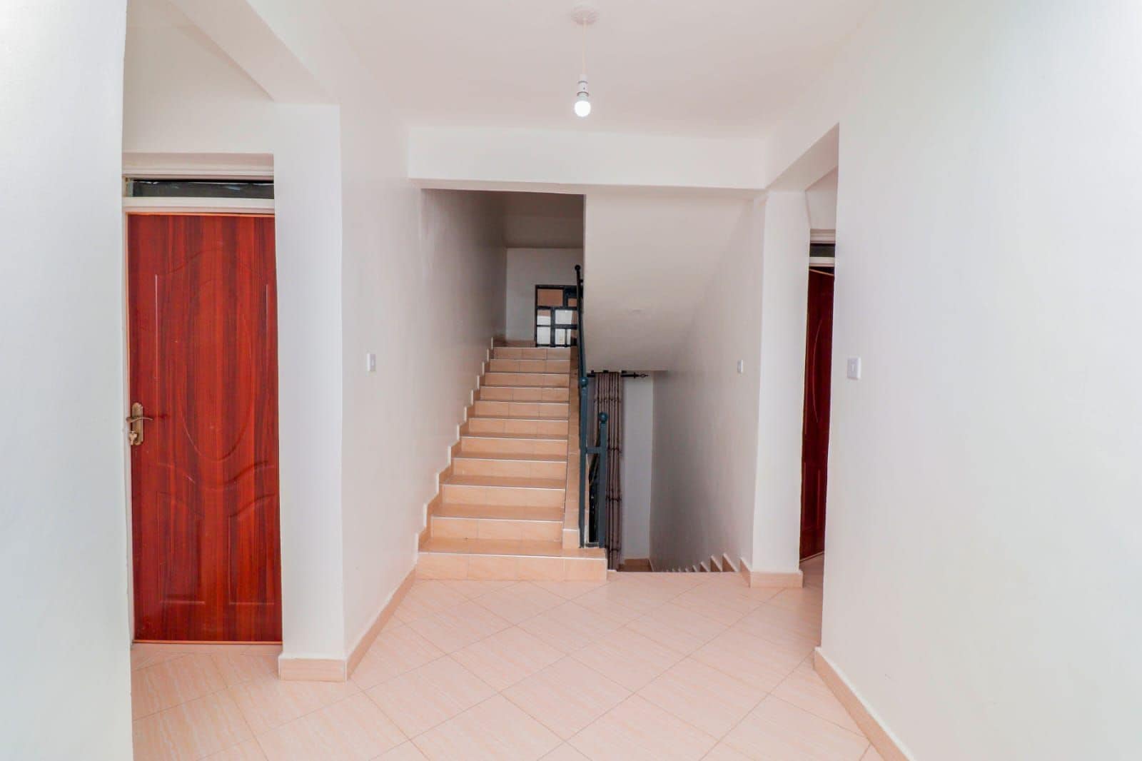 A hallway with a staircase leading to an upper floor, a red door on the left, and a white wall on the right.