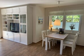 A dining area with a table and chairs, a television on a cabinet, and a window with a view of greenery. - Holiday home with 2 bedrooms (Greetsiel)