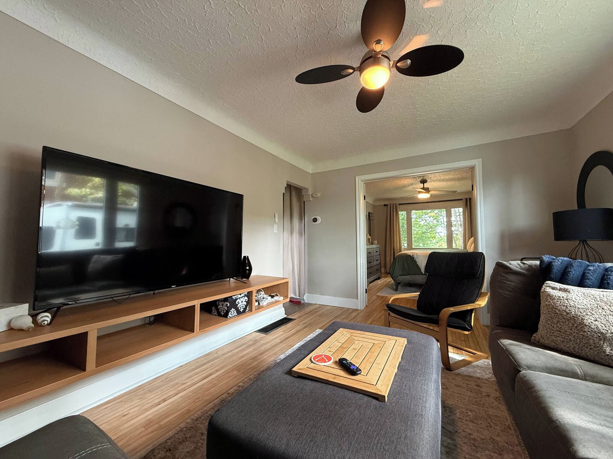 A modern living room with a flat-screen TV, wooden entertainment unit, a ceiling fan, and a cozy seating area with a sofa and ottoman.