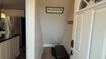 En smal gang fører til et køkken med hvide skabe og en sort bordplade. Et lille bord med en stol står op ad væggen, og et skilt over det siger 'MUDROOM'.