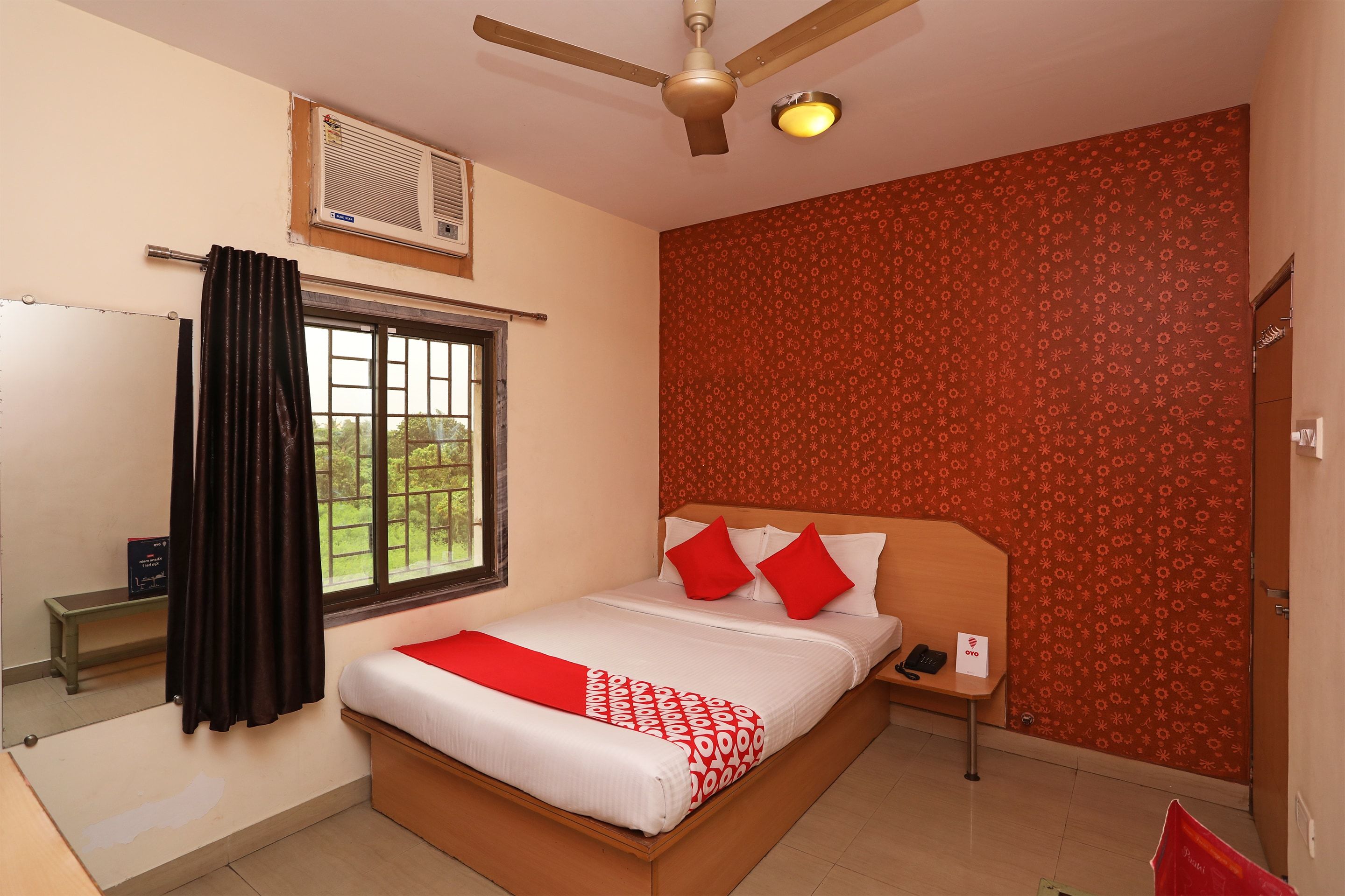 A hotel room with a bed, a nightstand, a telephone, a ceiling fan, a window with curtains, and a patterned wall.