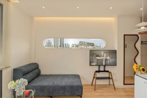 A modern living room with a grey sofa, a flat-screen TV mounted on the wall, and a cityscape view.