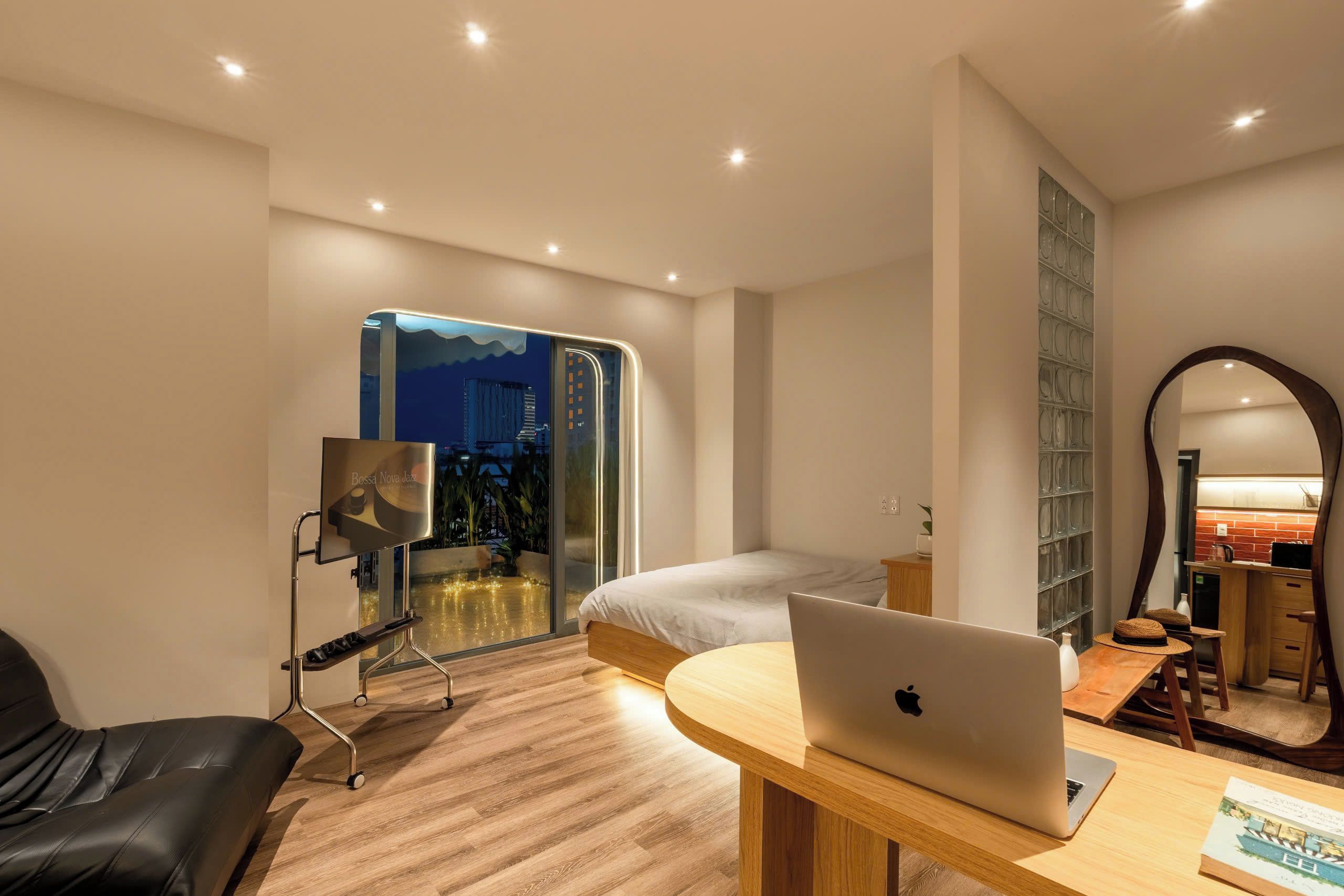 A modern bedroom with a bed, a desk with a laptop, a chair, a large mirror, and a view of the cityscape through a glass door.