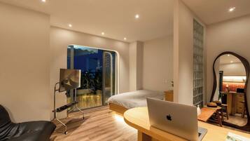 A modern bedroom with a bed, a desk with a laptop, a chair, a large mirror, and a view of the cityscape through a glass door.