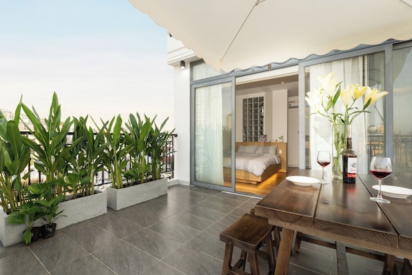 A modern balcony with potted plants, a dining table with chairs, and a view of a bedroom through a glass door.