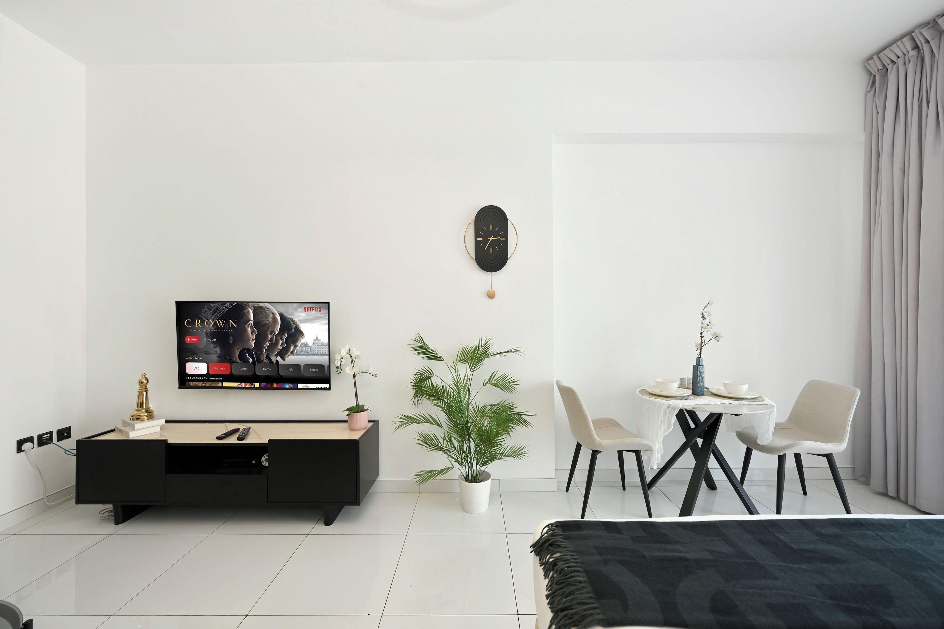 A modern living room with a flat-screen TV, a dining table with chairs, a potted plant, and a decorative clock on the wall.