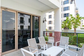 A balcony with white chairs and a table, a glass door leading to an indoor area, and a brick wall. - La Rive - Stylish 2 Bedroom Apartment (Dubai)