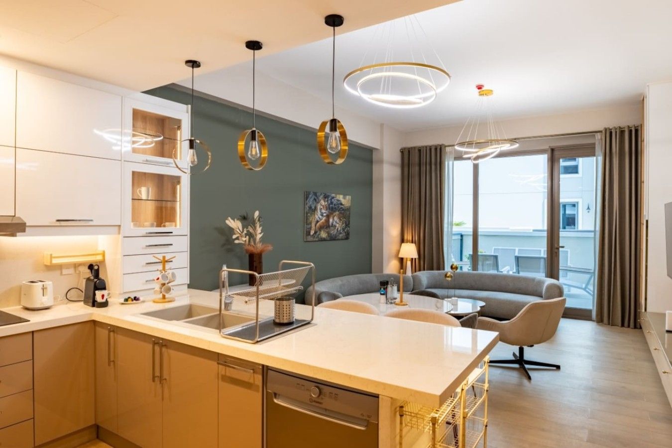 A modern hotel room with a kitchen area, a dining table, a sofa, and a large window with curtains.