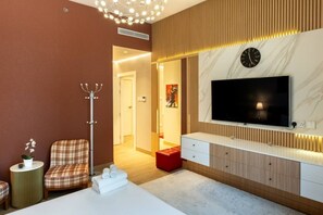 A modern living room with a flat-screen TV, a coat rack, a plaid sofa, and a red ottoman. - La Rive - Stylish 2 Bedroom Apartment (Dubai)