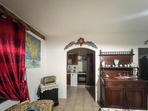 Una sala de estar con una cortina roja, un sillón estampado de flores, un aparador de madera con vajillas y una cocina visible a través de un arco.
