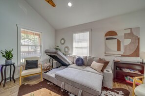Ein Zimmer mit einer Schlafcouch, zwei Fenstern mit Jalousien, einem kleinen Tisch mit einer Pflanze und einer Wand mit abstrakter Kunst.