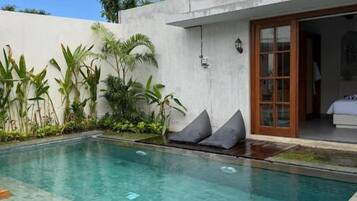 Una piscina con una silla de descanso en primer plano, rodeada de plantas y un edificio con puerta corredera de vidrio.