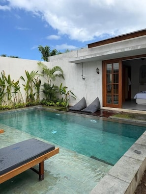 Piscina com espreguiçadeira em primeiro plano, cercada por plantas e um edifício com porta de correr de vidro.