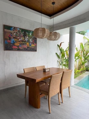 Uma sala de jantar com mesa de madeira e cadeiras de vime, luminária pendente e um quadro na parede.