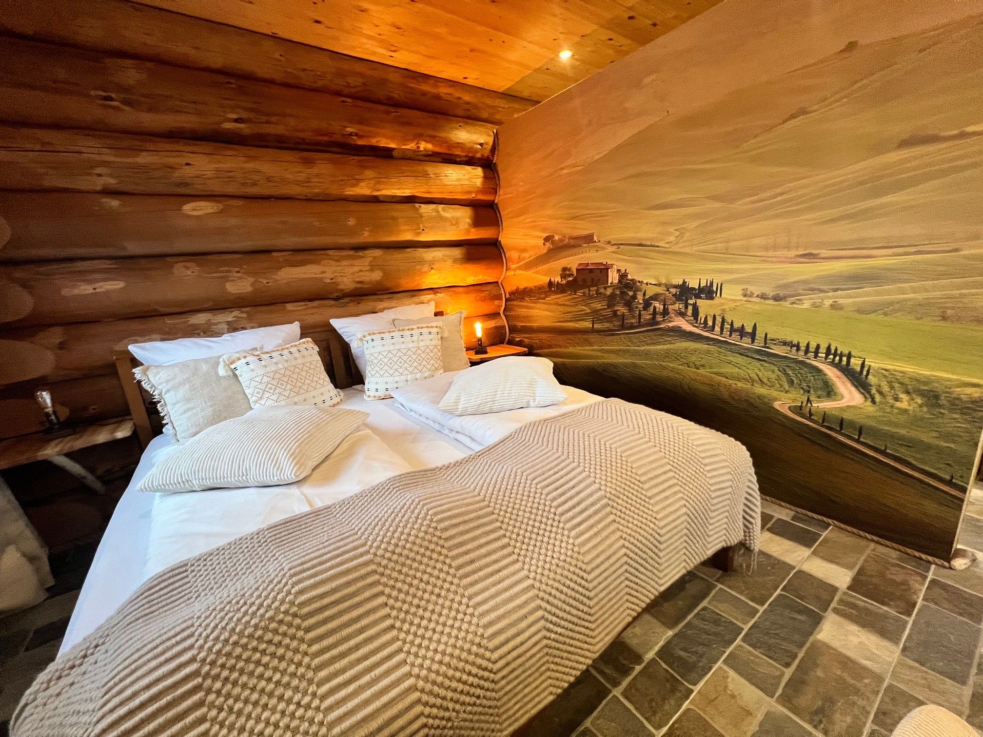 Une chambre à coucher avec un lit, des oreillers, une table de chevet et une grande fresque murale représentant un paysage.