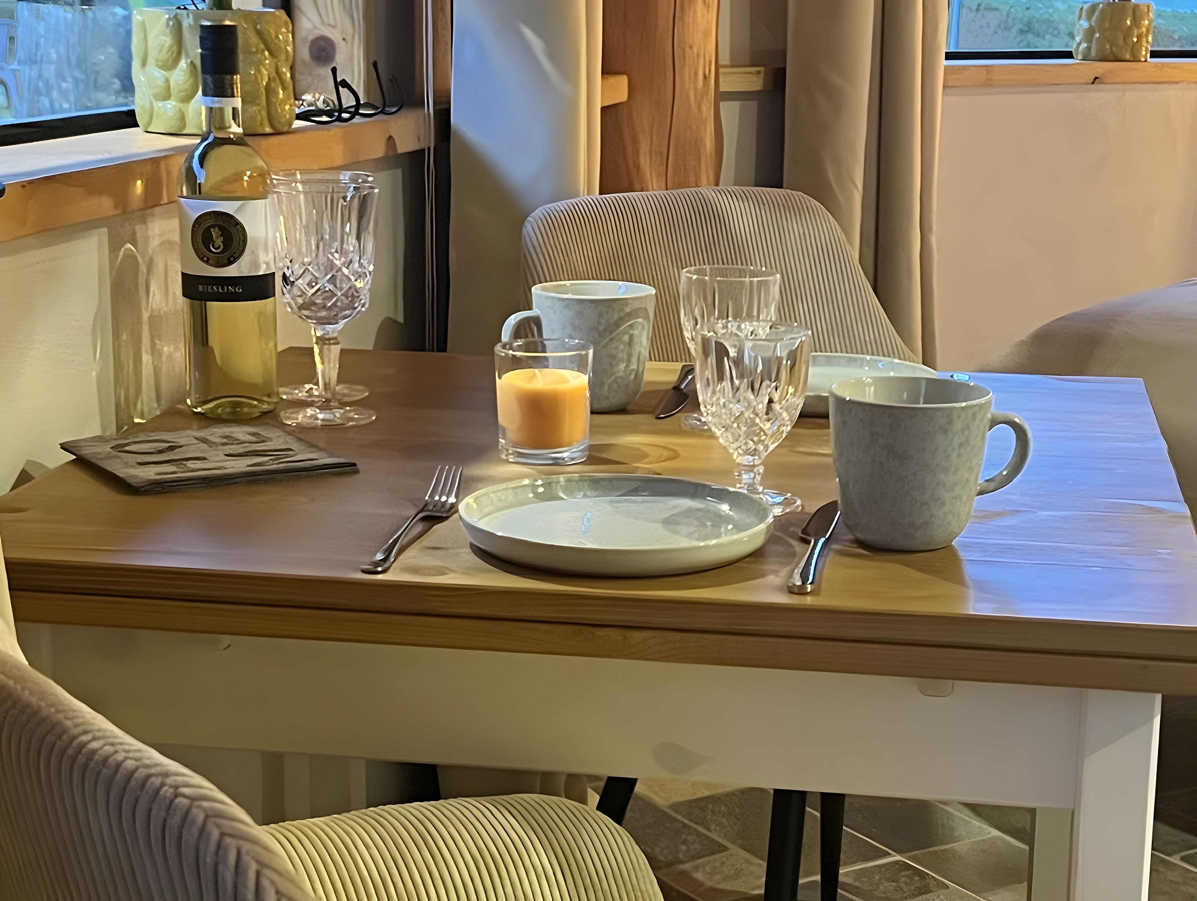Ein Esstisch gedeckt mit einer Flasche, Gläsern, einem Teller, einer Tasse und einer Kerze.