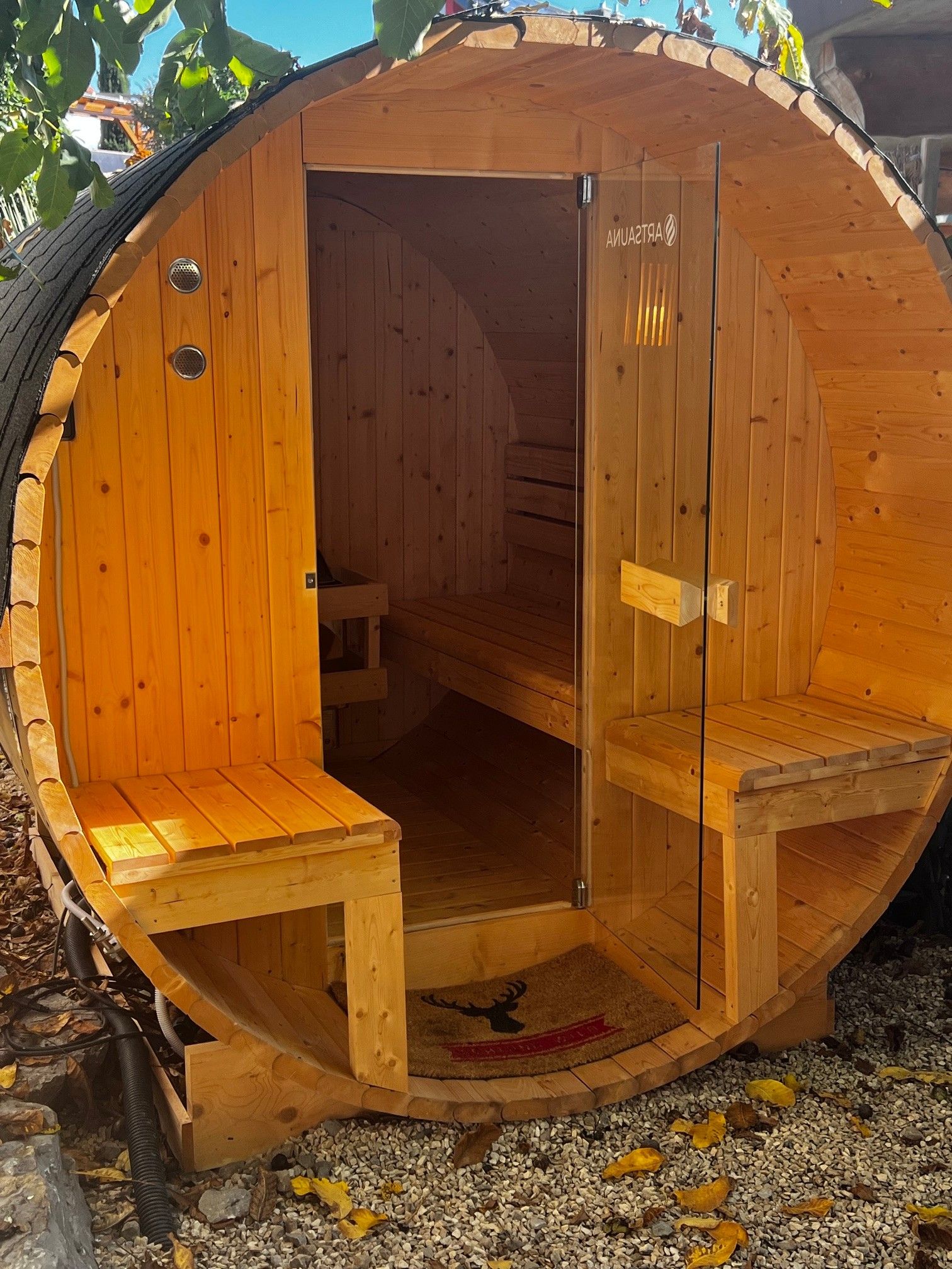 Eine Holzsauna mit Bänken und einer Tür, auf Kiesgrund, mit blauem Himmel.