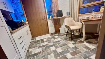 Une petite cuisine avec une porte en bois, un meuble blanc avec des tiroirs, une chaise beige et un sol carrelé.