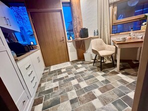 Une petite cuisine avec une porte en bois, un meuble blanc avec des tiroirs, une chaise beige et un sol carrelé.