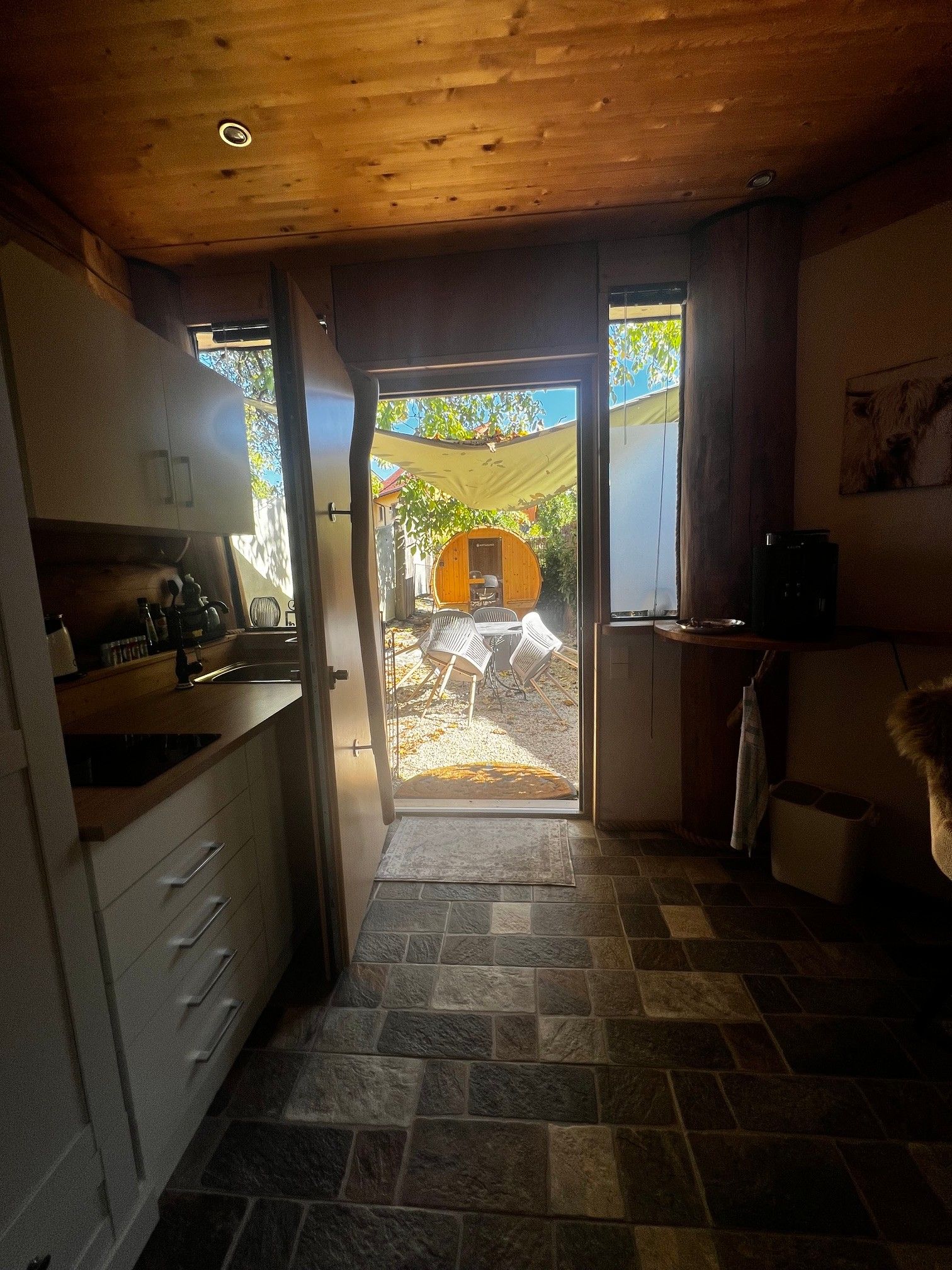 A kitchen with a stone floor, wooden ceiling, and a view of an outdoor seating area through a glass door.
