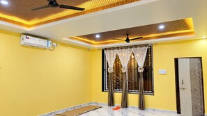 Una habitación con paredes amarillas, ventilador de techo, unidad de aire acondicionado y ventana con cortinas decorativas.
