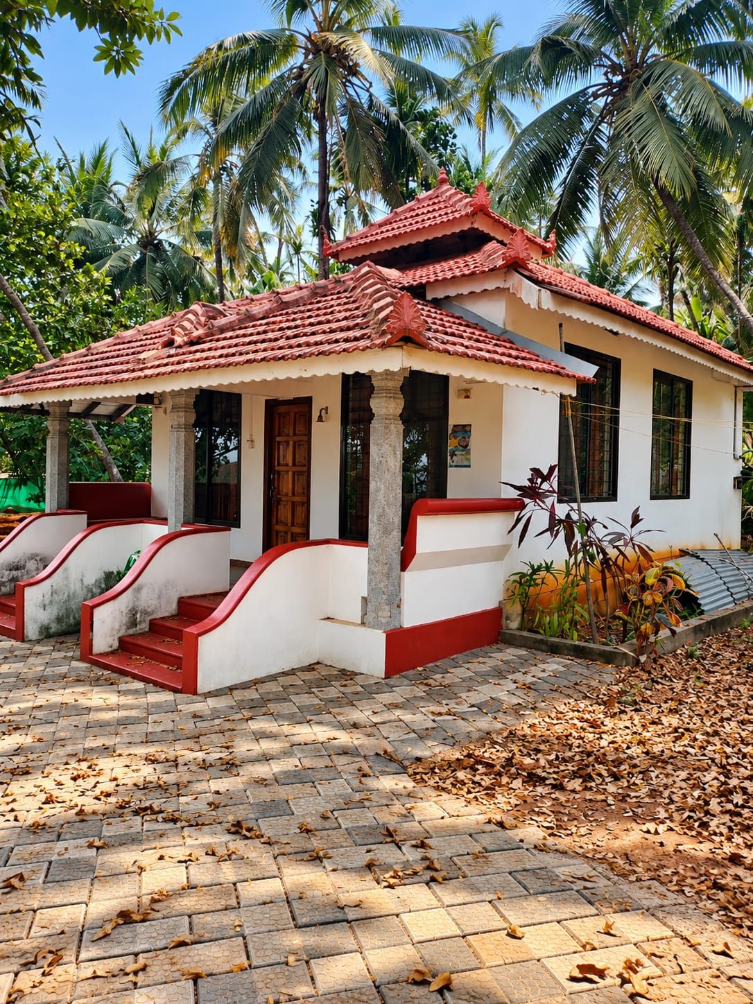 A traditional house with a tiled roof, white walls, and red accents, surrounded by palm trees and a paved area with fallen leaves.