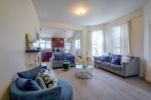 A spacious living area with a grey sofa, a blue armchair, and a coffee table. There is a kitchen in the background with dark wooden cabinets and a white microwave.