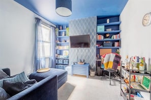 Ein Wohnzimmer mit einer Couch, einem Fernseher, einem Bücherregal und einem Schreibtisch.