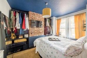 Een slaapkamer met een bakstenen muur, een bed met bloemenbeddengoed, een aan de muur bevestigde televisie en een kledingkast met hangende kleding.