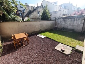 A small backyard with a wooden table and chairs, a grassy area, and a stone wall. - Le Bicoquet in Caen: An Elegant and Cozy Apartment (Caen)