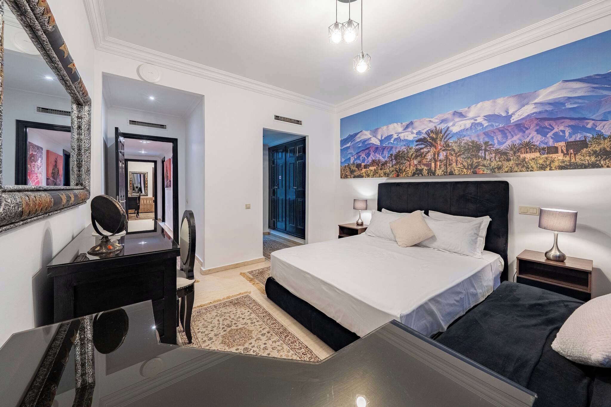 Ein Hotelzimmer mit einem großen Bett, einem Schreibtisch mit Spiegel, einem Nachttisch und einem Wandbild von einer Berglandschaft.