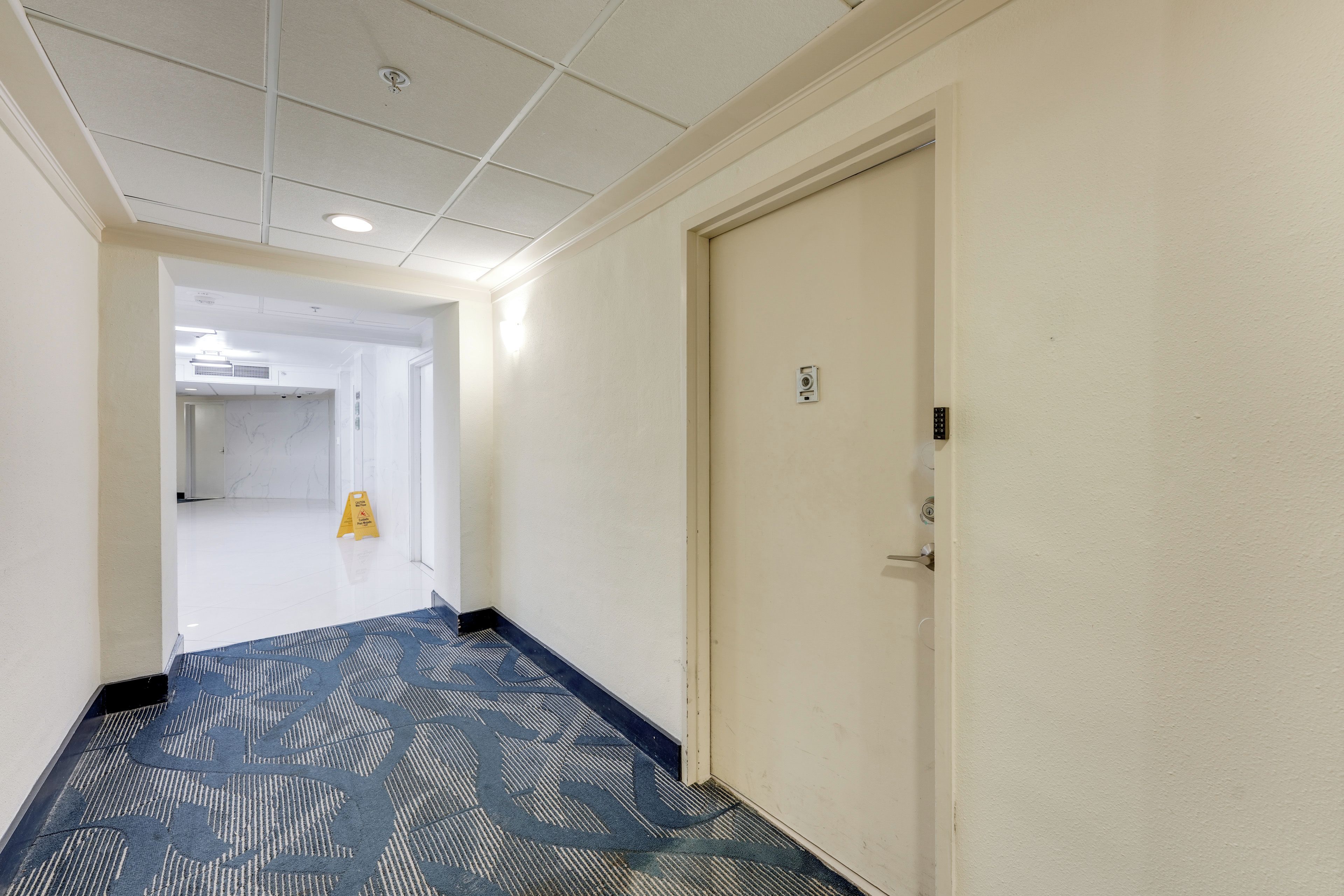 A hotel hallway with a carpeted floor, a door on the right, and a caution sign on the left.