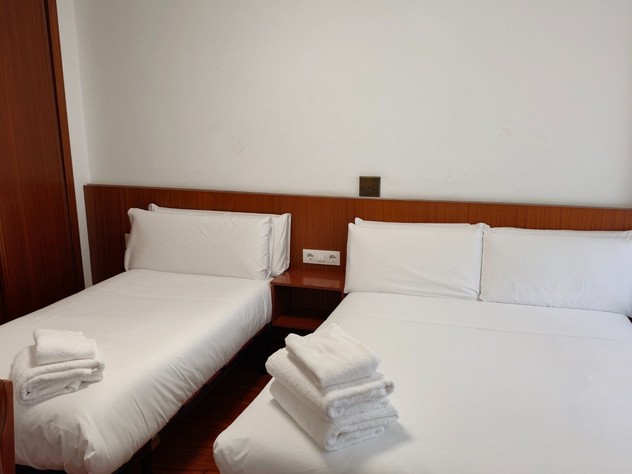Una habitación de hotel con dos camas individuales, cada una con ropa de cama y almohadas blancas, y un cabecero de madera. Hay una mesita de noche de madera entre las camas con un enchufe.