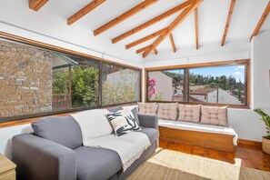 A room with a stone wall, a grey sofa, a bench with cushions, and a large window with a view of houses and greenery.