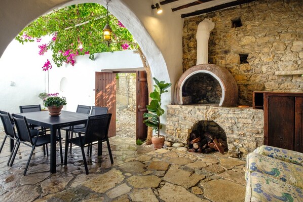 An outdoor patio with a stone table and chairs, a pizza oven, and a potted plant.