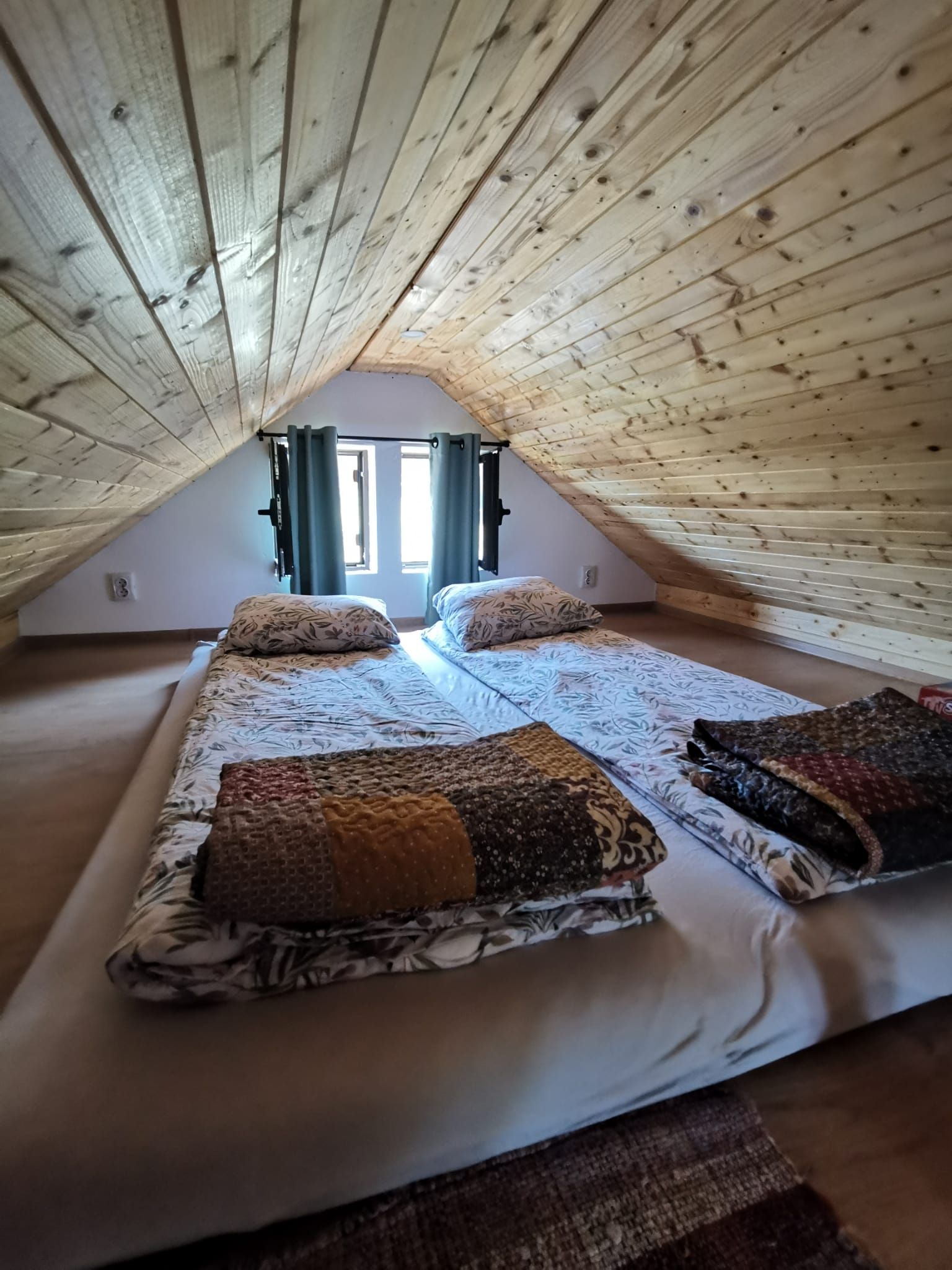 A loft bedroom with a sloped ceiling, two beds with patterned bedding, and a window with curtains.