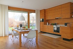 A modern kitchen with wooden cabinets, a dining table with chairs, and a view through large windows. - Holiday apartment with 2 bedrooms (Bodolz)