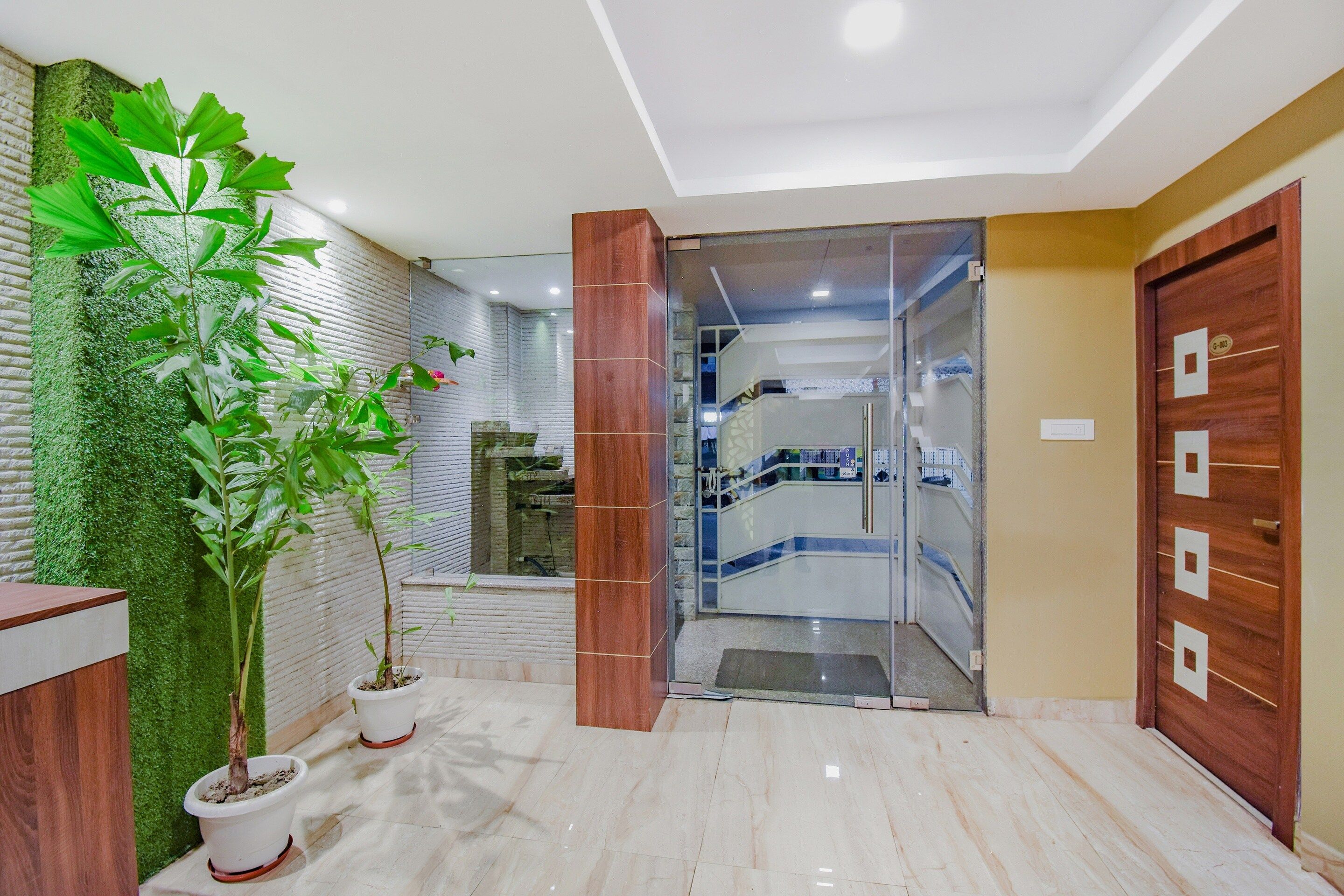 Un vestíbulo de hotel con mostrador de recepción, plantas en macetas, puerta de cristal de entrada y una escalera que conduce a pisos superiores.