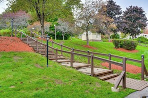 Eine Holztreppe führt einen sanften, grasbewachsenen Hang hinauf, im Hintergrund sind ein Zaun und Bäume zu sehen.