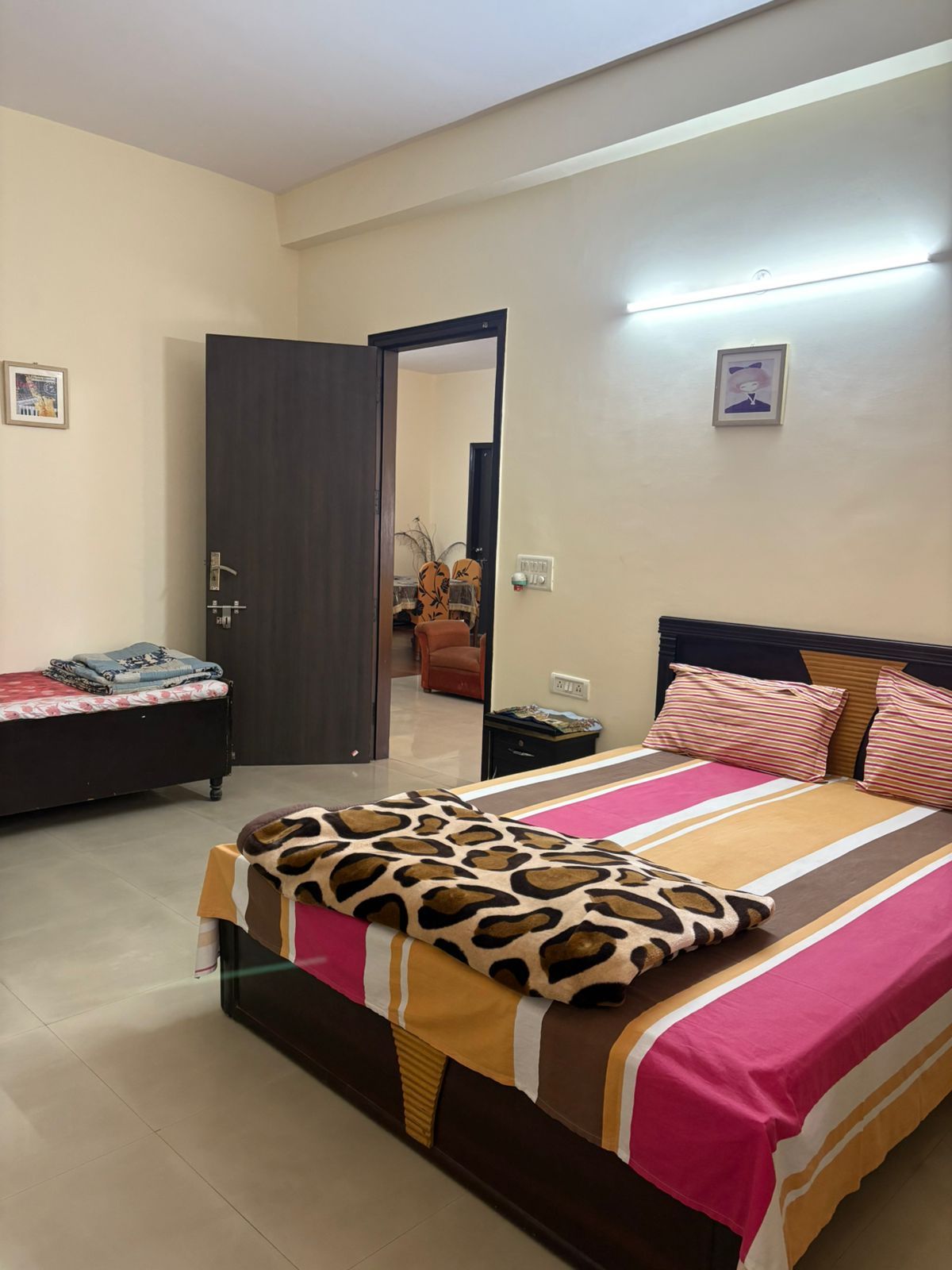 A bedroom with a bed, a nightstand, a door leading to another room, and a wall-mounted light fixture.