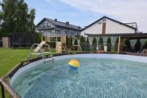 A swimming pool with a ladder and a ball, surrounded by a fenced yard with trees and a building in the background. - Baltic Harmony Villas (Grzybowo)
