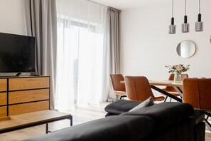 A modern living room with a sofa, a TV on a wooden cabinet, a dining table with chairs, and a large window with curtains. - Baltic Harmony Villas (Grzybowo)
