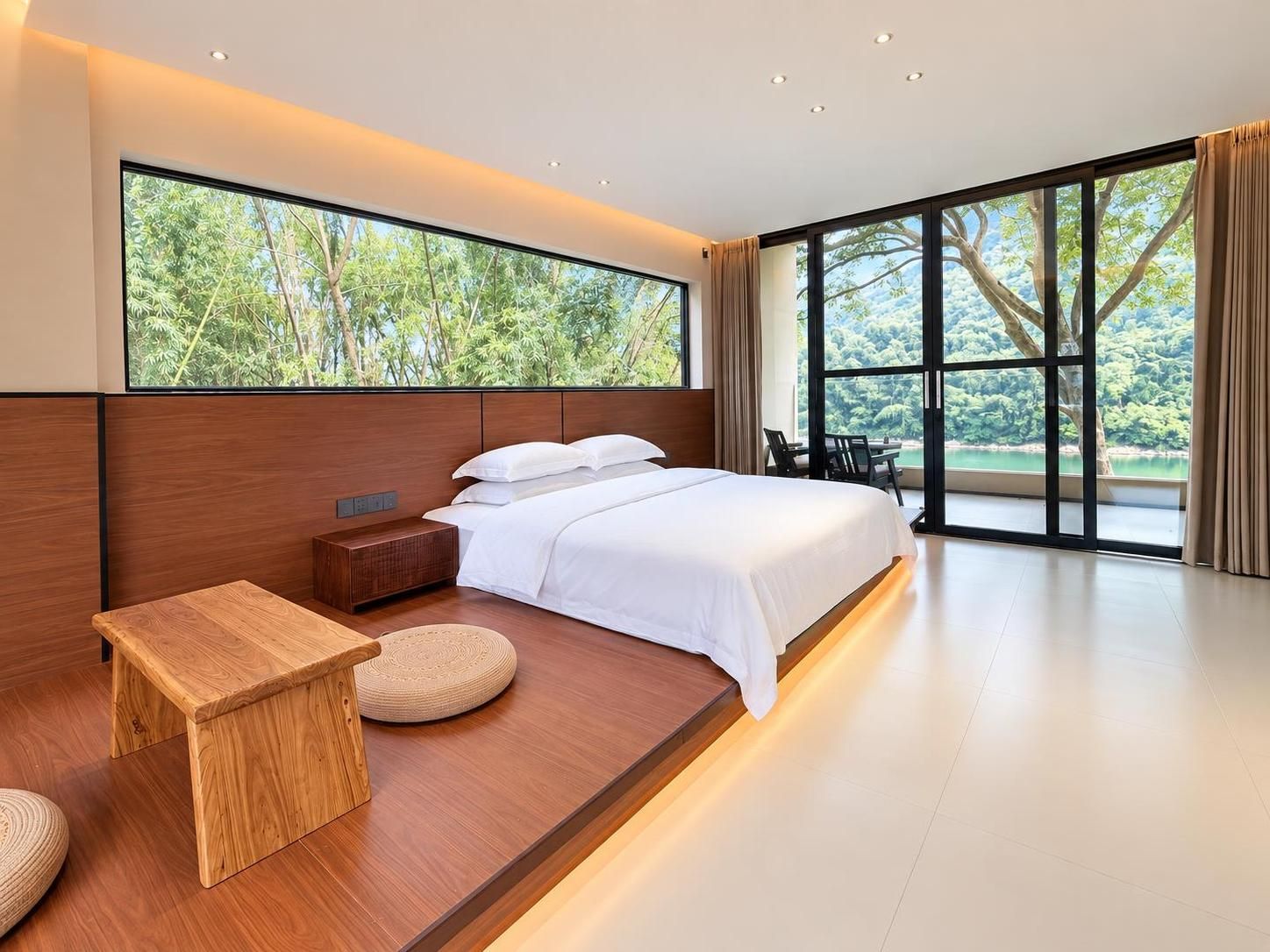 Un dormitorio moderno con una cama grande, una mesita de noche, un taburete pequeño de madera y vistas a zonas verdes a través de amplias ventanas.