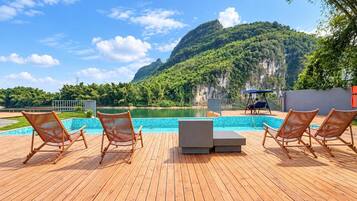 Una terraza de madera con piscina, sillas de descanso y vistas a un lago y montañas.