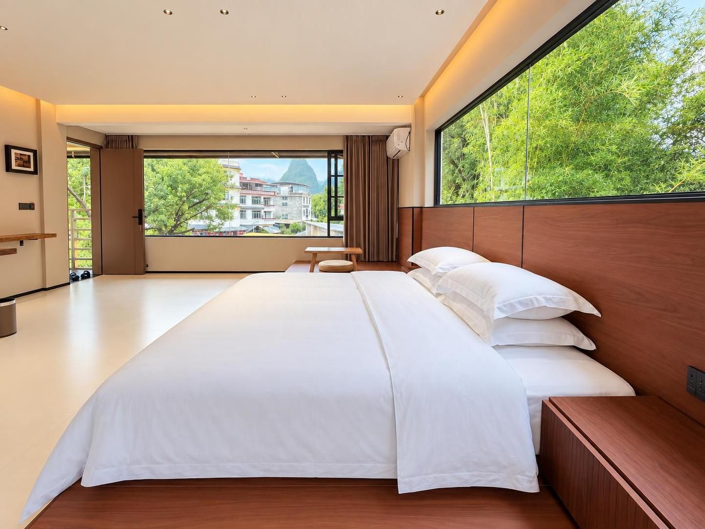 A hotel room with a large bed, a wooden headboard, a bedside table, a window with a view of buildings and trees, and a door leading outside.